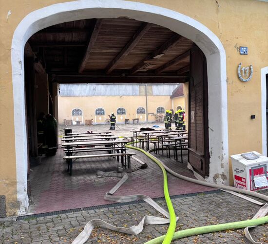 Ein Feuerwehrschlauch führt durch ein offenes Tor zu einem Innenhof, wo Feuerwehrleute nahe Tischen stehen.