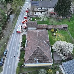 Luftaufnahme eines Hauses mit Feuerwehr und Einsatzkräften, die an einem Notfall arbeiten, umgeben von blühenden Bäumen.