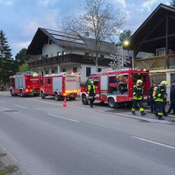 Feuerwehrleute arbeiten an einem Einsatz vor einem Gebäude, während Feuerwehrfahrzeuge bereitstehen.