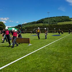 Menschen in Feuerwehruniformen tragen Holzbretter auf einem sonnigen Sportplatz während eines Wettkampfs.