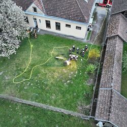 Feuerwehrleute arbeiten im Garten eines Hauses, während ein Schlauch verlegt ist und ein Baum blüht.