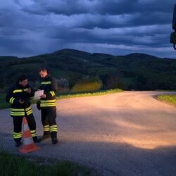 Zwei Feuerwehrleute sprechen über Funk neben einem orangefarbenen Warnkegel auf einer Straße in der Abenddämmerung.
