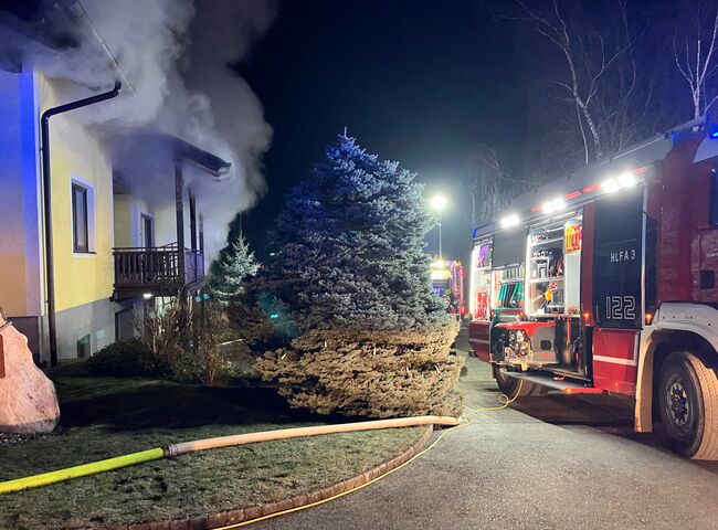 Ein Feuerwehrauto steht vor einem rauchenden Haus, während Löschschläuche im nächtlichen Einsatz bereitliegen.